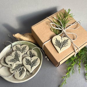 May include: A collection of off-white ceramic heart ornaments featuring a black botanical fern print. The hearts are suspended by thin white string. One heart is tied to a square brown cardboard gift box, while others rest in a shallow bowl.