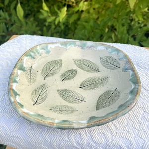 Può includere: Un piatto in ceramica rotondo e poco profondo con smalto bianco sporco maculato. L'interno presenta diverse impronte di foglie verde scuro con venature visibili. Il bordo ondulato e irregolare è rifinito con smalto azzurro e marrone.