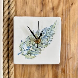 Peut inclure: Horloge murale carrée blanche avec un motif de feuille de fougère dans des tons verts et bleus. L'horloge a des aiguilles noires et un centre doré. Elle est fixée sur une surface en bois.