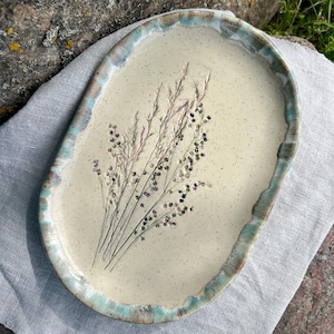 Può includere: Un vassoio ovale in ceramica con superficie color crema maculata, caratterizzato da un'impronta botanica di erbe essiccate e piccoli fiori. Il vassoio presenta un bordo rialzato e ondulato smaltato nei toni del blu, verde e marrone.
