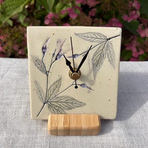 May include: A square ceramic clock with a cream-coloured speckled surface featuring a dark blue botanical illustration of leaves and buds. The clock has three black hands centred on the face and rests on a rectangular light wood stand.