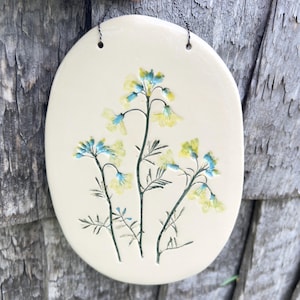 Op de afbeelding: Een ovaalvormige keramische wandplaat met een gladde crèmekleurige glazuurlaag. Het oppervlak bevat een ingedrukte botanische illustratie van wilde bloemen met gele bloemblaadjes en blauwgroene stengels en bladeren, opgehangen aan een dunne gedraaide metalen draad.