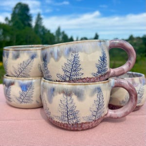 Puede incluir: Seis tazas de cerámica apiladas, con una base color crema con diseños de hojas de helecho azules y asas moradas. Los bordes tienen un esmalte azul y la base una banda marrón rojiza. Las tazas están sobre una superficie rosa, con un fondo verde.