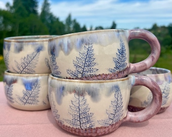 May include: A stack of six ceramic mugs with a cream-colored base, blue fern leaf design, and purple handles. The rims have a blue glaze, and the bottom has a reddish-brown band. The mugs are stacked on a pink surface with a green background.