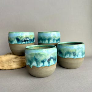 Puede incluir: Cuatro tazas de cerámica hechas a mano con un esmalte único. Las tazas tienen una base marrón claro y están cubiertas con una mezcla de esmalte azul y verde, creando un efecto marino. Las tazas son redondas y tienen un acabado liso.