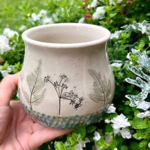 May include: A cream-colored ceramic mug featuring dark botanical imprints of ferns and wildflowers on the exterior. The base of the mug is decorated with a textured pattern of small, rounded blue-green indentations encircling the bottom.
