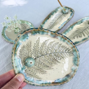 Può includere: Un set di piatti in ceramica fatti a mano con centro color crema e bordo smerlato blu-verde. Ogni pezzo presenta un'impronta dettagliata di foglia di felce nera. Un piatto ovale include una perlina in ceramica per bastoncini d'incenso.