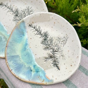 May include: Two ceramic plates with a speckled, off-white surface. One plate has a blue and turquoise glazed edge. Each plate features a gray botanical design. The plates are on a striped cloth.