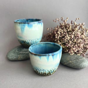 Op de afbeelding: Twee handgemaakte keramische bekers met een blauw-witte glazuur. De bekers hebben een getextureerde basis en worden gepresenteerd op grijze stenen, met gedroogde bloemen op de achtergrond. De binnenkant van de bekers is helderblauw.