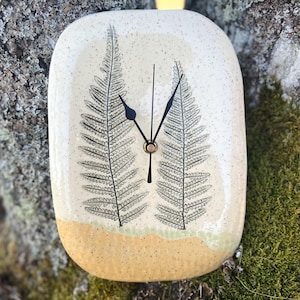 May include: An oval ceramic wall clock with a speckled cream and yellow glaze. The clock face features two detailed black fern leaf illustrations and three thin black clock hands centered on the surface. The edges are rounded and smooth.