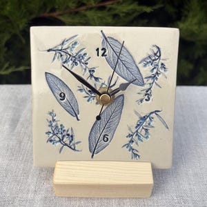 May include: A square ceramic clock with a cream-coloured surface featuring blue botanical leaf and sprig imprints. The clock face displays black numbers at the 12, 3, 6, and 9 positions, with black hour, minute, and second hands. It rests on a light wood stand.