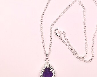 Op de afbeelding: Zilverkleurige ketting met een druppelvormige hanger. De hanger heeft een dieppaarse steen, omringd door kleine, heldere stenen. De ketting is delicaat en heeft een karabijnsluiting.