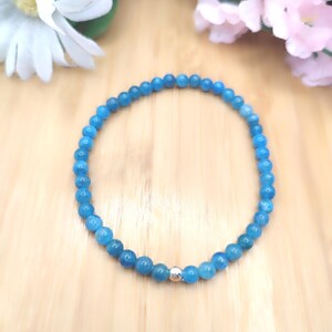 May include: A blue beaded bracelet with a silver accent bead. The bracelet is made of small, round, light blue beads. The bracelet is displayed on a light brown wooden surface, with flowers in the background.