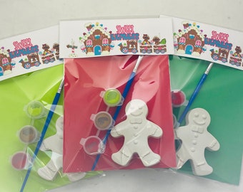 May include: Three gingerbread house themed paint your own gingerbread man kits. Each kit includes a white ceramic gingerbread man, a paintbrush, and four small pots of paint. The paint colors are red, green, brown, and yellow.