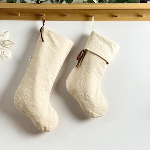 Op de afbeelding: Twee crèmekleurige kerstsokken hangen aan een houten plank. De sokken hebben een textuur en bruine lussen. Een papieren ster en groen zijn ook zichtbaar.