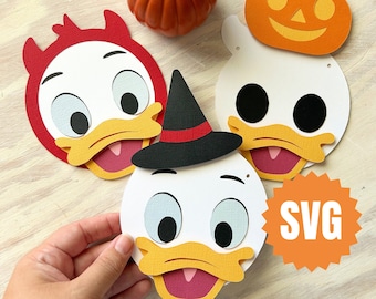 Może przedstawiać: Cztery papierowe wycięcia kreskówkowej kaczki w różnych kostiumach na Halloween. Kostiumy obejmują kapelusz czarownicy, rogi diabła, prześcieradło ducha i dynię. Wycięcia znajdują się na drewnianej powierzchni z małą pomarańczową dynią.