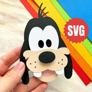 May include: A papercraft cutout of Goofy's face. The cutout is made of black, white, brown, and pink paper. The cutout is held in a hand against a rainbow background. The word "SVG" is in a red starburst.