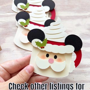 May include: Papercraft Santa mug with black Mickey Mouse ears, white whipped cream, and a red handle. The Santa face has a pink nose and a white beard. The mug is decorated with green holly and red berries. Text: Check other listings for a Mickey-eared version!