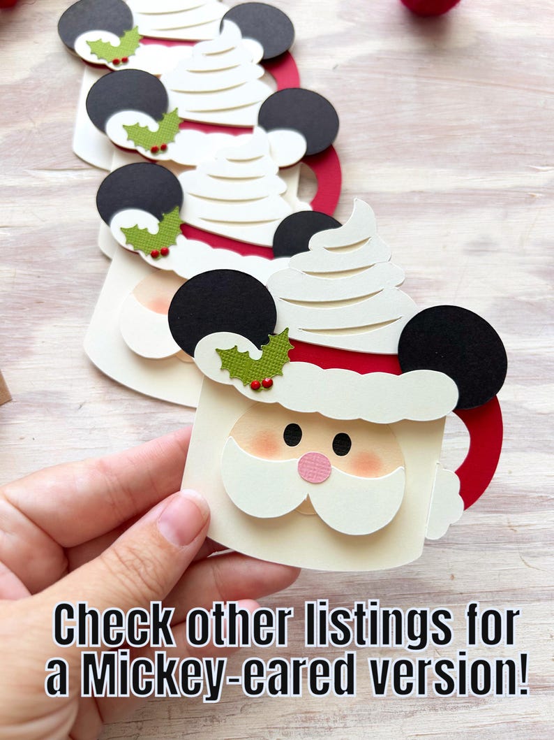 May include: Papercraft Santa mug with black Mickey Mouse ears, white whipped cream, and a red handle. The Santa face has a pink nose and a white beard. The mug is decorated with green holly and red berries. Text: Check other listings for a Mickey-eared version!