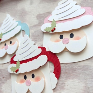 May include: Close-up of three Christmas-themed paper cutouts. Each cutout features a Santa Claus face with a white beard and hat, a pink nose, and black eyes. The Santa faces are on mugs with red, blue, and white handles. White whipped cream tops the mugs.