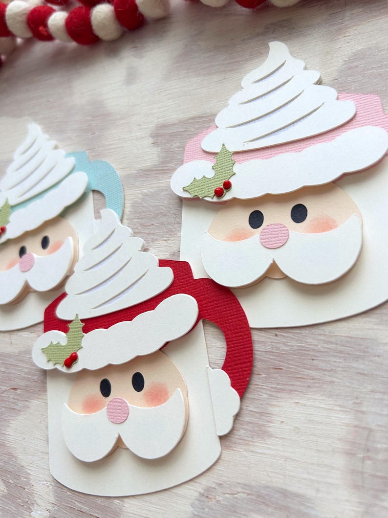 May include: Close-up of three Christmas-themed paper cutouts. Each cutout features a Santa Claus face with a white beard and hat, a pink nose, and black eyes. The Santa faces are on mugs with red, blue, and white handles. White whipped cream tops the mugs.