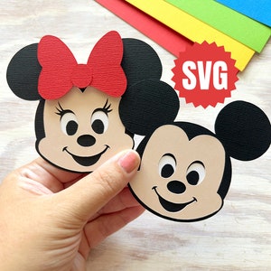 May include: Two paper cutouts of Mickey Mouse and Minnie Mouse. Mickey Mouse is brown with black ears and a black nose. Minnie Mouse is brown with black ears and a red bow. The cutouts are on a wooden surface with the word "SVG" in a red starburst.