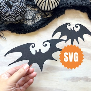 May include: Two black paper bats with white eyes and a swirl design. The bats are on a white wooden surface with a bright orange circle that says "SVG".