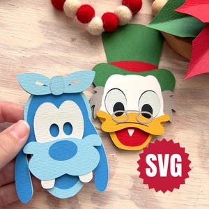 Puede incluir: Dos recortes de papel de personajes de dibujos animados, uno azul con un lazo y el otro con un sombrero verde y gafas. También se ven una guirnalda roja y blanca y un logotipo SVG rojo.