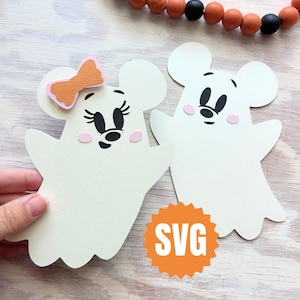 May include: Two white ghost-shaped cutouts with mouse ears and faces, one with a pink and orange bow. The ghost cutouts have black eyes and noses, and pink cheeks. An orange SVG logo is visible. A decorative beaded garland is in the background.