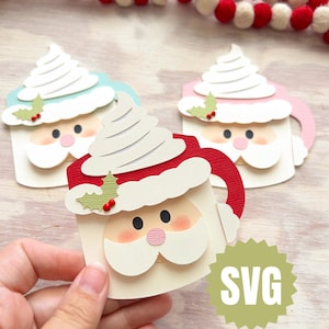 May include: Three Santa-themed paper cutouts shaped like mugs, each topped with whipped cream. The mugs are in red, pink, and blue, with white beards and rosy cheeks. A green SVG logo is visible. A festive garland with red and white balls is in the background.