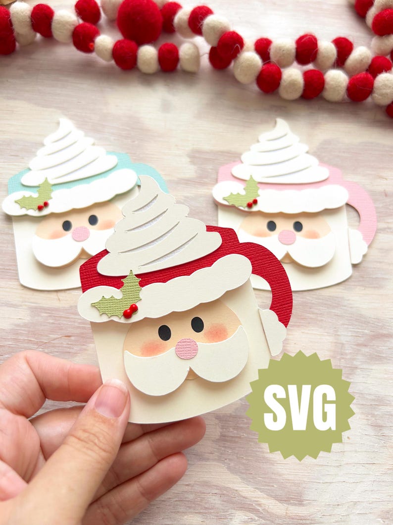 May include: Three Santa-themed paper cutouts shaped like mugs, each topped with whipped cream. The mugs are in red, pink, and blue, with white beards and rosy cheeks. A green SVG logo is visible. A festive garland with red and white balls is in the background.