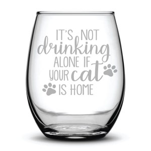 May include: A clear glass wine glass with the text "It's not drinking alone if your cat is home" etched on the side. The text is in a cursive font and there are two paw prints on either side of the text.