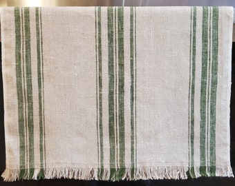 May include: A rectangular kitchen towel made of natural beige fabric featuring vertical dark green stripes of varying widths. The bottom edge of the towel is finished with a decorative fringe that includes both beige and green threads.