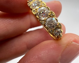 May include: A gold ring with five large, clear diamonds set in a row. Smaller diamonds are set between the larger stones. The ring is held between fingers against a white background, showcasing the ring's sparkle and design.