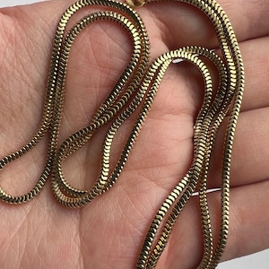 Könnte beinhalten: Eine goldfarbene Schlangenkette. Die Kette besteht aus kleinen, ineinandergreifenden Gliedern, die eine glatte, flexible Textur erzeugen. Die Halskette wird auf einer Hand präsentiert, um ihre Länge und ihr Design zu zeigen. Der Verschluss ist oben sichtbar.