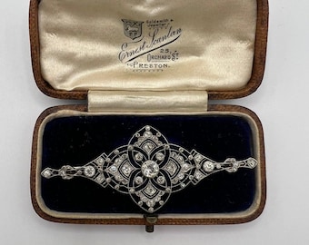 Könnte beinhalten: Antike silberne Brosche mit klaren Edelsteinen, präsentiert in einer Vintage-Schmuckschatulle. Die Brosche hat ein zentrales Blumendesign und ist auf einem dunkelblauen Samtinnenraum platziert. Die Schatulle ist braun mit einem cremefarbenen Satinfutter.