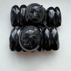 May include: Two black plastic bracelets with a carved design of a woman's head in a helmet. The bracelets are made of multiple stacked pieces.