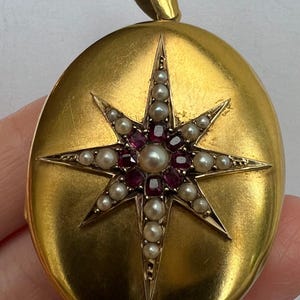 Peut inclure: Pendentif en forme de médaillon ovale doré avec un motif d'étoile. L'étoile est ornée d'une perle centrale entourée de pierres rouges et de petites perles. Le pendentif a une boucle pour une chaîne.