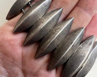 May include: A silver-toned bracelet featuring six elongated, leaf-shaped links. The links are connected by small metal bars and have a textured surface. The bracelet is shown resting on a person's palm.