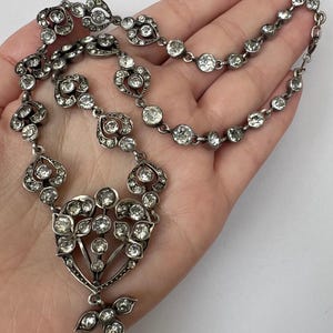 Può includere: Una collana argentata con pietre preziose trasparenti. La collana presenta motivi floreali e a forma di cuore, con una catena delicata. Le pietre sono rotonde e a goccia, incastonate in una montatura dettagliata e ornata.