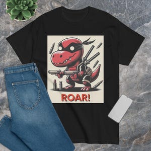 Könnte beinhalten: Schwarzes T-Shirt mit einem Aufdruck eines roten Dinosauriers, der eine schwarze Maske trägt und eine Pistole hält. Der Dinosaurier ist von Kugeln umgeben und der Text "ROAR!" ist darunter gedruckt.