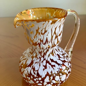May include: A small amber glass pitcher with a white speckled design. The pitcher has a rounded body and a curved handle.