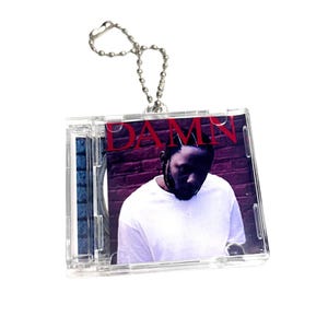 May include: A clear plastic keychain with a silver chain. The keychain features a miniature CD case with the album cover for Kendrick Lamar's "DAMN." album. The album cover features Kendrick Lamar in a white shirt against a red brick wall.