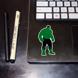 Può includere: Un adesivo verde e nero di Hulk, un supereroe immaginario, in piedi con i pugni serrati. L'adesivo è su un laptop nero.