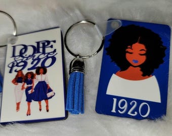 May include: Two blue and white keychains with a tassel. One keychain features three women in blue and white outfits with the text "Dope Since 1920". The other keychain features a black woman with blue lips and the text "1920".