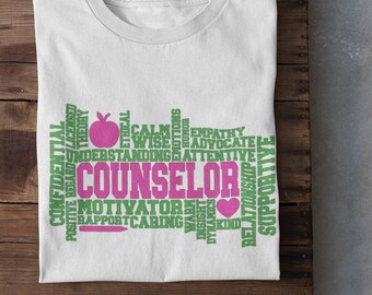 May include: White t-shirt featuring the word "COUNSELOR" in pink and green, with associated terms such as "MOTIVATOR" and "EMPATHY." The design also incorporates an apple and a heart. The t-shirt is folded on a wooden surface.