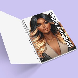 May include: A spiral bound notebook featuring a black and white illustration of a woman with long blonde and black hair. The cover of the notebook reads "MONTHLY GOALS" in white text.