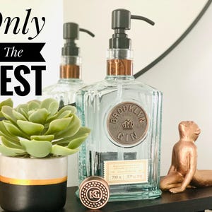 Könnte beinhalten: Klare Brooklyn Gin-Flasche aus Glas mit schwarzem Pumpenspender und Kupferakzenten. Eine kleine Bronzestatue eines Affen und eine Sukkulentenpflanze in einem schwarz-weiß-goldenen Topf sind ebenfalls zu sehen. Der Text "Only The BEST" steht oben.