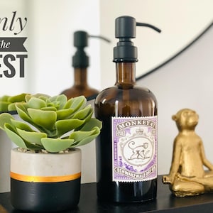 Könnte beinhalten: Eine braune Glasflasche mit einem schwarzen Pumpspender, gefüllt mit Flüssigkeit. Die Flasche trägt ein Etikett mit der Aufschrift "Monkey 47 Schwarzwald Dry Gin". Die Flasche steht auf einer schwarzen Oberfläche neben einer goldenen Affenfigur und einer Sukkulente im Topf.