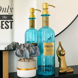 Könnte beinhalten: Zwei blaue Glasflaschen mit goldenen Pumpen und Etiketten. Die Flasche rechts trägt den Text "ITALICUS ROSOLIO DI BERGAMOTTO" auf dem Etikett. Die Flaschen stehen auf einer Holzoberfläche mit einer kleinen Sukkulentenpflanze im Topf und einer goldenen Affenfigur.